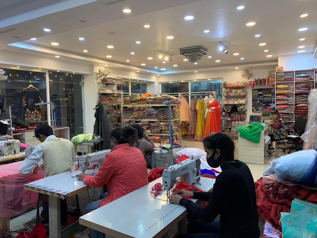 The workroom at Adaa Bazaar: tailors at sewing machines, with bolts of fabric and finished pieces on the shelves behind them.
